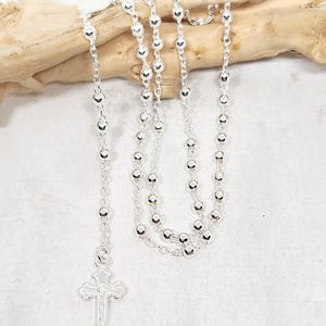 Può includere: Una collana rosario d'argento con perline rotonde e un pendente a croce dettagliato. La collana è drappeggiata su un pezzo di legno alla deriva su uno sfondo chiaro. Le perline sono distanziate uniformemente e riflettono la luce.
