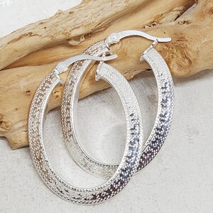 May include: A pair of silver oval hoop earrings. The earrings have a textured pattern along the outer edge. The earrings are displayed on a light brown wooden surface.