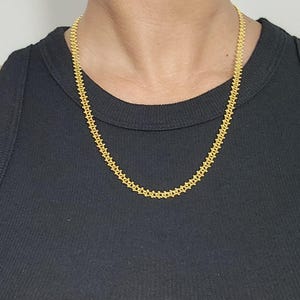Pu&ograve; includere: Una collana a catena d'oro con un design delicato e intricato. La catena &egrave; indossata da una persona con una camicia nera.