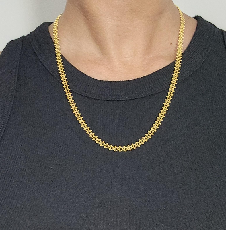 Pu&ograve; includere: Una collana a catena d'oro con un design delicato e intricato. La catena &egrave; indossata da una persona con una camicia nera.