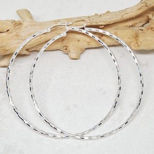 Puede incluir: Un par de pendientes de aro grandes y plateados. Los pendientes tienen un diseño geométrico texturizado y un cierre de bisagra. Los aros se muestran sobre un fondo neutro con un trozo de madera flotante.