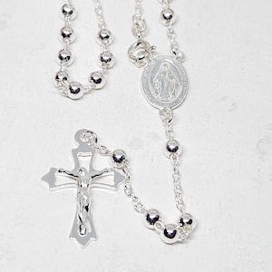May include: A silver rosary with round beads and a crucifix. The rosary features a detailed crucifix with a figure of Jesus and an oval pendant with an image of the Virgin Mary.