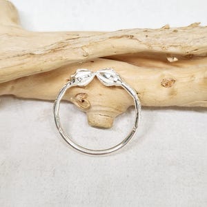 May include: A silver-colored ring with a floral design. The ring features two flower bud shapes at the top, connected to a smooth, rounded band. The ring is displayed on a light-colored, textured surface.