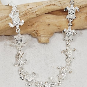 May include: A silver-colored necklace featuring multiple turtle charms. The charms are connected by delicate links, creating a charming and whimsical design. The necklace is displayed on a wooden surface.