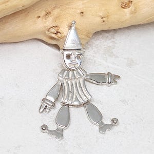 May include: A silver charm in the shape of a jester with a pointed hat, a striped shirt, and bells on its shoes.