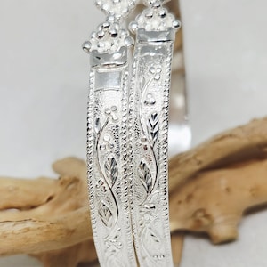 May include: Two silver bangles with intricate floral and leaf patterns. The bangles have a detailed design with a textured surface. The top of the bangles have a decorative floral design. The bangles are displayed on a wooden branch.