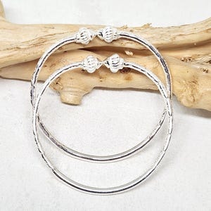May include: Two silver bangles with textured bands and decorative ball accents. The bangles are displayed on a light-colored surface with a piece of driftwood in the background. The jewelry is simple and elegant.
