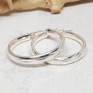 May include: A pair of silver hoop earrings. The earrings are round with a smooth, polished finish and a subtle engraved design. The hoops are connected with a hinged clasp. The earrings are displayed on a light-colored surface.