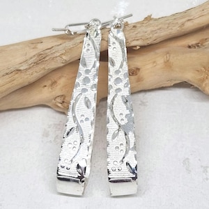 May include: A pair of long, silver-toned drop earrings with a tapered, flat design. The surface of each earring features a textured pattern of small circles and curved leaf-like shapes, with a polished finish that reflects light across the metal.