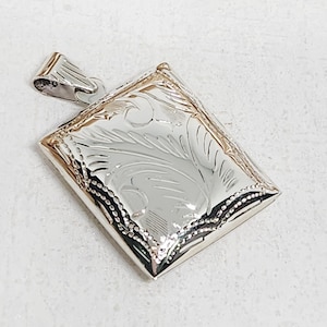 Könnte beinhalten: Ein rechteckiger Silberanhänger mit einem eingravierten floralen Design. Der Anhänger hat einen dekorativen Rand und eine Schlaufe für eine Kette. Das Metall hat eine polierte Oberfläche, die das Licht reflektiert.