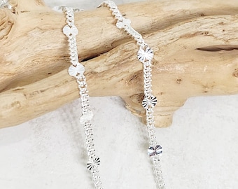 May include: A silver necklace with a delicate chain design. The necklace features small, circular, and flower-shaped accents. The jewellery is displayed on a piece of driftwood against a white background.