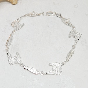 Pode incluir: Uma pulseira de prata com berloques em forma de ilha. A pulseira tem uma corrente delicada e um fecho seguro. Os berloques são representações em miniatura de ilhas, com designs e texturas intrincados. É uma joia.