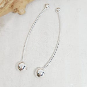 May include: A pair of silver-colored, curved dangle earrings. Each earring features a small, round ball at the top and a larger, round ball at the bottom, connected by a thin, curved bar. The earrings have a minimalist, modern design.