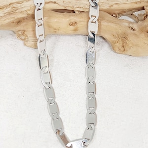 Puede incluir: Un collar plateado con un diseño de eslabones rectangulares repetidos. El collar se muestra sobre un fondo claro con un trozo de madera flotante.
