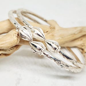 Può includere: Due braccialetti d'argento con un design floreale. I braccialetti hanno una superficie dettagliata e strutturata e sono adornati con elementi floreali stilizzati. I gioielli sono esposti su un pezzo di legno alla deriva su sfondo bianco.