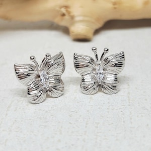 May include: A pair of silver butterfly stud earrings. Each earring features detailed wing patterns and a central, clear gemstone. The earrings are designed with a post-back closure for secure wear. The butterfly design adds a touch of nature-inspired elegance.