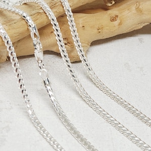 Könnte beinhalten: Nahaufnahme mehrerer silberfarbener Kettenhalsketten. Die Ketten haben ein gewebtes Muster und werden auf einer hellen Oberfläche mit einem Holzstück im Hintergrund gezeigt. Die Ketten sind reflektierend und scheinen aus Metall zu sein.