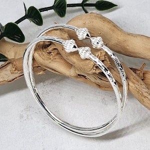 May include: Two silver bangles with a textured design and decorative accents. The bangles are stacked on top of each other.