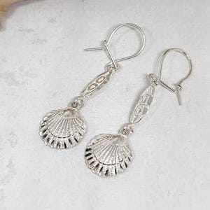 May include: A pair of silver-toned drop earrings featuring a textured scallop shell charm suspended from a decorative, elongated filigree link. The earrings are attached to simple silver-toned ear wires, showcasing a metallic finish and detailed shell patterns.