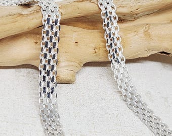 Puede incluir: Un collar de cadena de tono plateado con un diseño de malla tejida ancho y plano. Los eslabones metálicos están dispuestos en un patrón de rejilla repetitivo, creando una estructura flexible similar a una cinta que se apoya sobre madera.