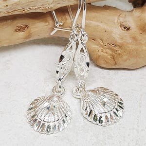 May include: A pair of silver-toned drop earrings featuring a textured seashell charm suspended from a decorative, elongated, openwork metal link. The earrings are attached to curved ear wires, showcasing a polished, reflective finish throughout.