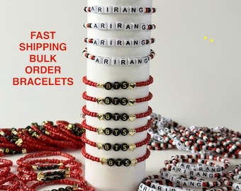May include: A collection of beaded bracelets featuring white square beads with black letters spelling ARIRANG, and red beaded bracelets with black circular beads spelling BTS accompanied by small gold-toned heart-shaped charms.
