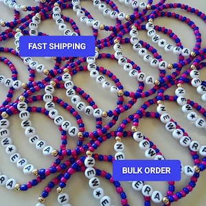 May include: A collection of beaded bracelets with the text "NEW ERA" spelled out in white letter beads. The bracelets feature alternating pink and blue beads, with gold-colored accents. The bracelets are scattered across a light-colored surface.
