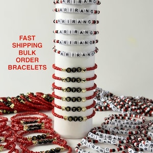 May include: A collection of beaded bracelets featuring white beads with black letters spelling ARIRANG and black beads with gold letters spelling BTS alongside small gold heart charms, with red and white seed beads on elastic cord.