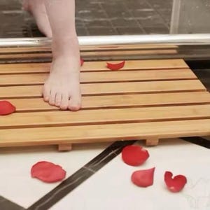 Pode incluir: Um tapete de banho de bambu natural com um design de ripas. O tapete está colocado sobre um piso de azulejos com pétalas de rosa vermelhas espalhadas ao redor.