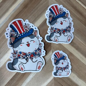 May include: Three stickers featuring a cartoon cat wearing a patriotic top hat and a floral wreath. The cat is white with gray spots and a smiling face. The hat has red and white stripes and blue stars.