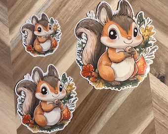 Puede incluir: Tres pegatinas de ardillas de dibujos animados. Las ardillas son marrones, naranjas y blancas, sosteniendo nueces y rodeadas de hojas de otoño. Las pegatinas varían en tamaño y están colocadas sobre una superficie de madera.