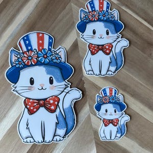 Puede incluir: Tres pegatinas de gatos en diferentes tamaños. Cada gato es blanco con detalles azules, con una pajarita roja y un sombrero de copa azul decorado con estrellas y rayas rojas, blancas y azules. Los sombreros también están adornados con flores.