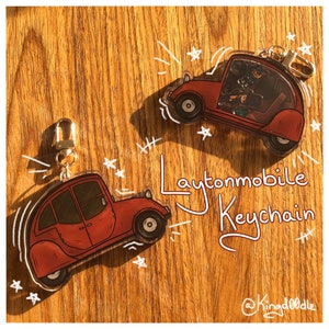 Puede incluir: Dos llaveros Laytonmobile rojos con cierres dorados se muestran sobre una superficie de madera. Los llaveros presentan un diseño de coche de dibujos animados con el texto "Laytonmobile Keychain" en una fuente manuscrita. Acentos de estrellas y líneas blancas rodean los llaveros.