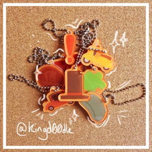 Puede incluir: Un conjunto de llaveros coloridos con un sombrero, un signo de exclamación, un coche y otros diseños. Los llaveros están unidos a una cadena plateada y se muestran sobre un fondo de corcho. El texto "@Kingdoodle" está escrito en el corcho.