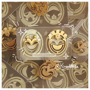 Op de afbeelding: Goudkleurige metalen spelden met een gestileerd maskerontwerp. De spelden zijn verpakt op kaarten met de tekst "MIRACLE MASK SET PINS". De spelden hebben een glanzende afwerking en zijn in een groep gerangschikt.