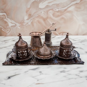 May include: A set of ornate copper Turkish coffee cups and a matching pot with a grinder on a marble surface.