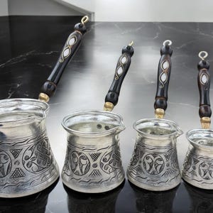 May include: Four silver Turkish coffee pots with intricate engraved designs and wooden handles with white accents.