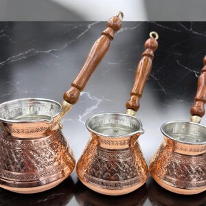 May include: Three copper Turkish coffee pots with wooden handles. The pots are decorated with an embossed pattern and have a shiny finish.