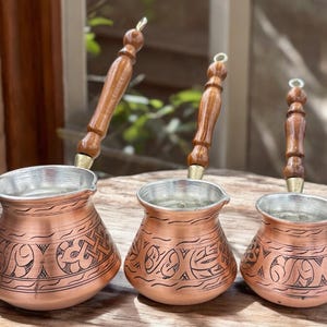 May include: Three copper cezve coffee pots with wooden handles. The pots have a decorative etched design and a silver-colored interior. The handles are a light brown color. The cezves are displayed on a wooden surface.