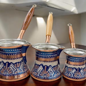 May include: Three copper Turkish coffee pots with blue and white enamel designs and wooden handles. The pots are of different sizes.