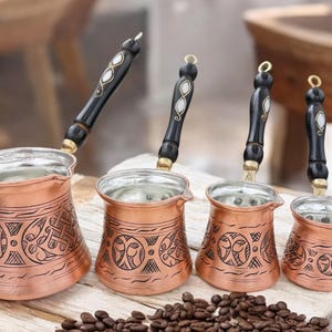 May include: Four copper Turkish coffee pots with black wooden handles and intricate engraved designs. The pots are arranged in order of size, with the largest pot in the front and the smallest pot in the back. The pots are sitting on a wooden surface with coffee beans scattered around them.