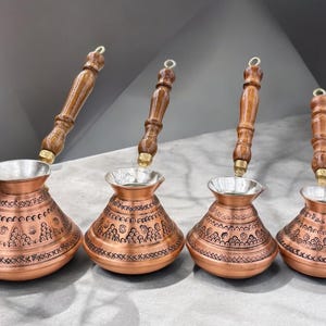 以下が含まれることがあります： 木製の取っ手が付いた銅製のトルコ式コーヒーポットが4つ。ポットは複雑な模様で飾られており、銀色の裏地が付いています。