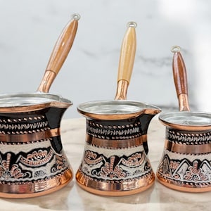 Può includere: Tre caffettiere turche in rame con manici in legno. Le caffettiere sono decorate con intricati disegni in bianco e nero e hanno una finitura in rame e argento.