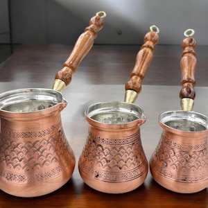 May include: Three copper coffee pots with wooden handles. The pots have a decorative pattern and are arranged in order of size.