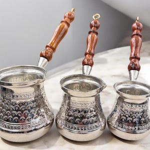 May include: Three silver Turkish coffee pots with wooden handles. The pots have an ornate design and are arranged in a row from largest to smallest.