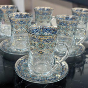 Op de afbeelding: Set van zes heldere glazen theekopjes met ingewikkelde blauwe en gouden patronen. Elk kopje wordt geleverd met een bijpassend schoteltje.
