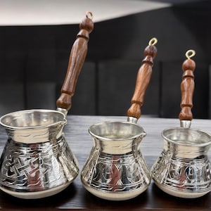 May include: Three silver Turkish coffee pots with wooden handles. The pots are decorated with intricate patterns and are of different sizes.