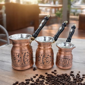May include: Three copper Turkish coffee pots with intricate floral designs and wooden handles. The pots are arranged in order of size, with the largest pot in the front and the smallest pot in the back. The pots are sitting on a wooden table with coffee beans scattered around them.