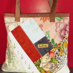 Pode incluir: Uma bolsa tote de patchwork com al&ccedil;as de couro marrom. A bolsa &eacute; feita de diferentes tecidos com padr&otilde;es florais, um tecido vermelho e branco e um tecido azul e branco. A bolsa tem uma etiqueta que diz "ScobCr&eacute;a".