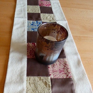 Peut inclure: Un chemin de table blanc avec un motif patchwork brun, beige et rouge. Le chemin de table est décoré d'un bougeoir en verre brun.